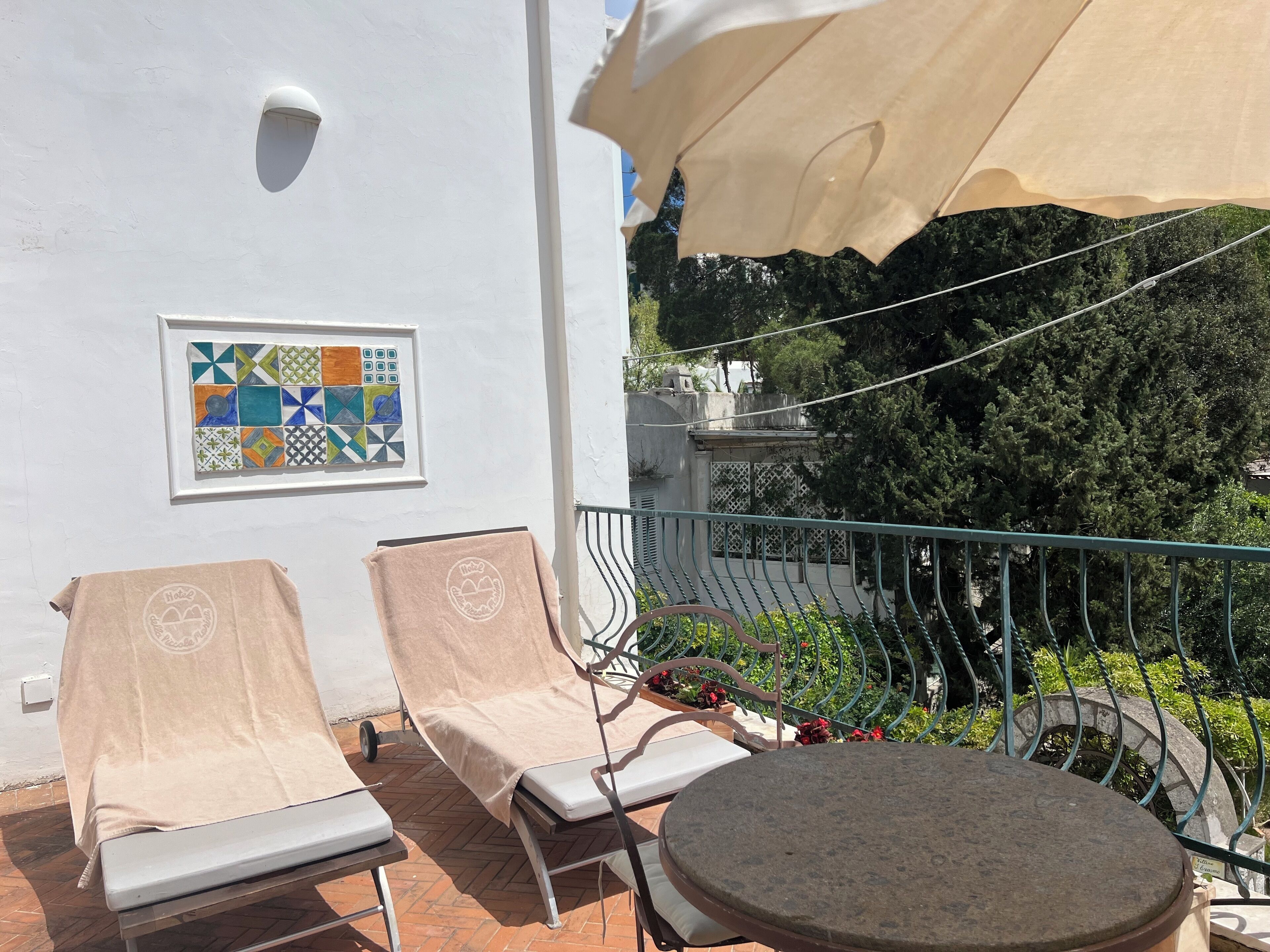 A balcony with two sun loungers and a table, a framed mosaic on the wall, and a view of trees and buildings in the background.