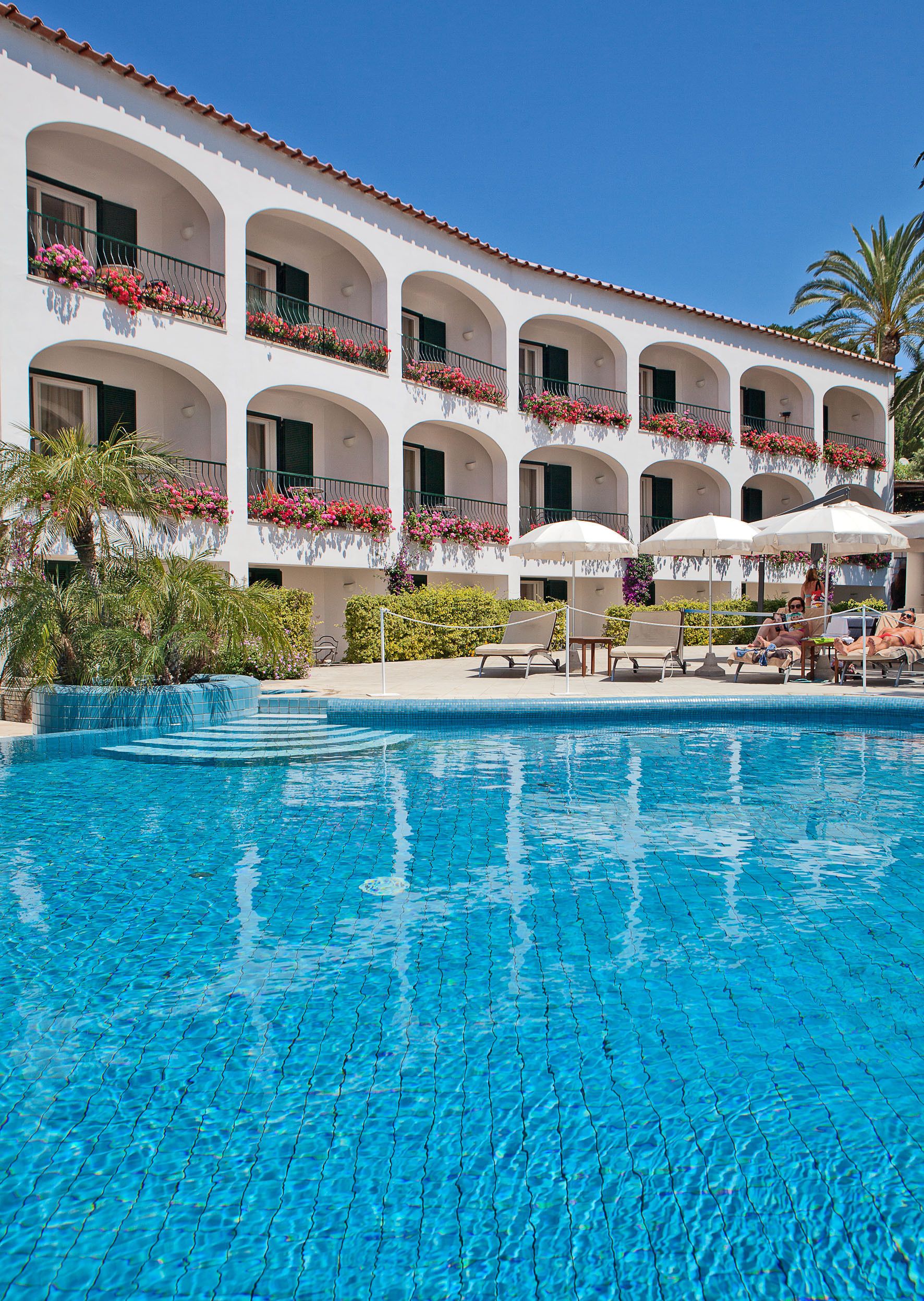 Un hotel con piscina, varios balcones con macetas de flores y zonas de estar al aire libre.