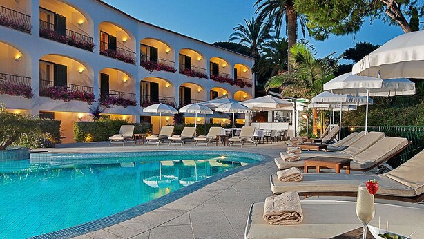 Zona de piscina con sillones, sombrillas y un edificio con balcones.