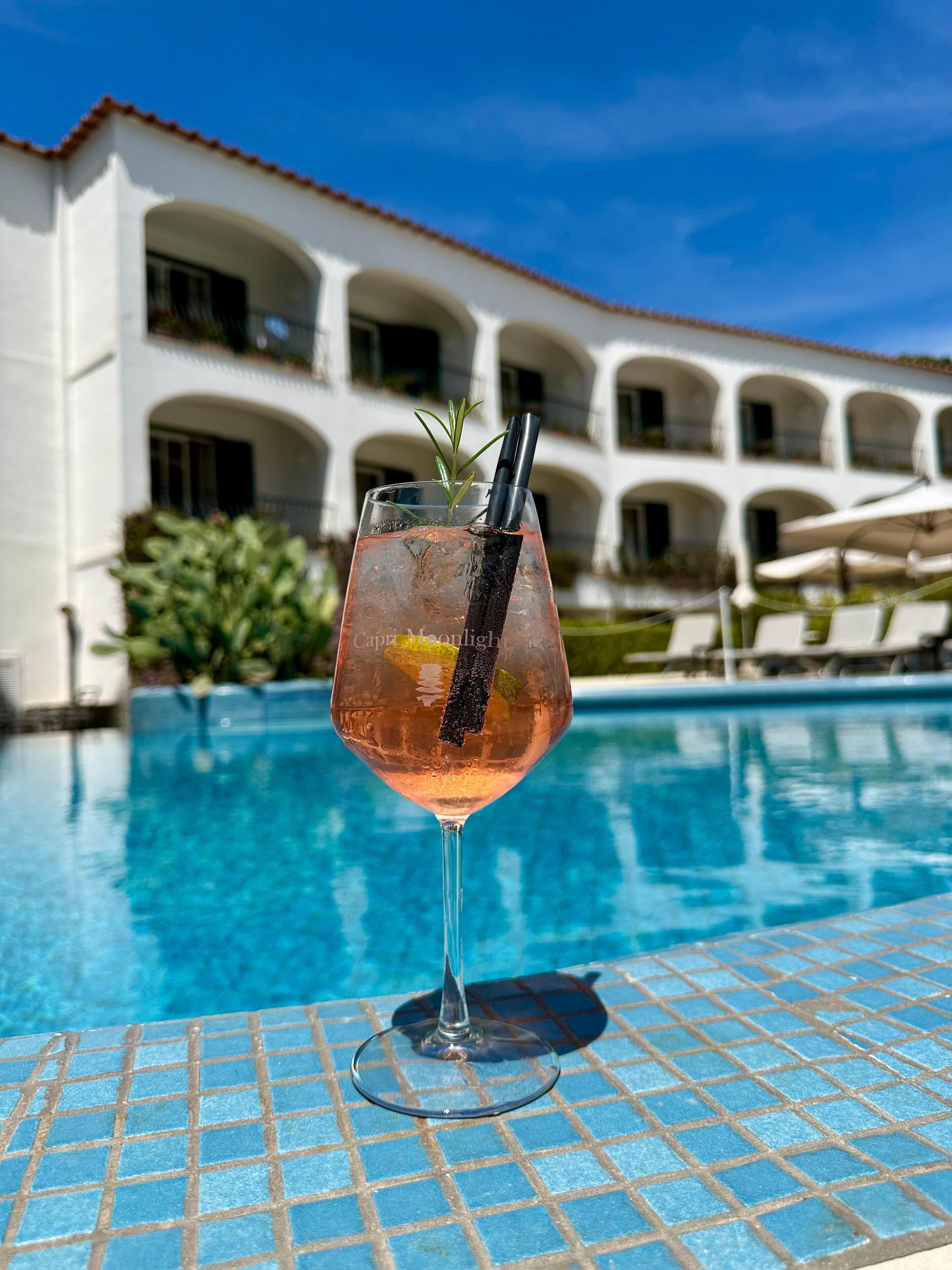 Ein Glas rosa Cocktail mit Rosmarinzweig und schwarzem Strohhalm auf einem gefliesten Tisch am Pool, im Hintergrund ein weißes Gebäude mit Bögen.