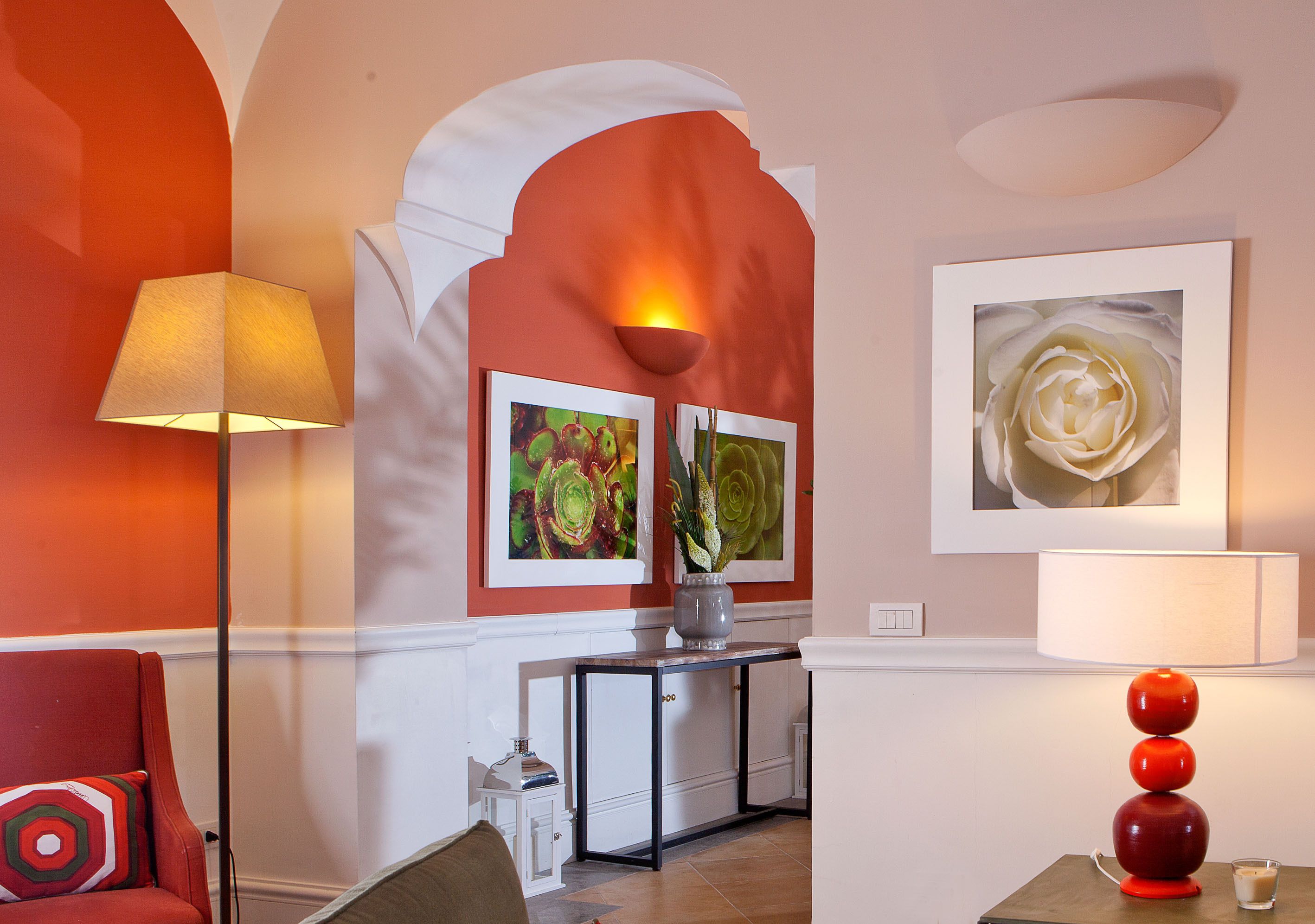 A hotel lobby with a red armchair, a standing lamp, a console table with a vase, and framed floral artwork on the walls.