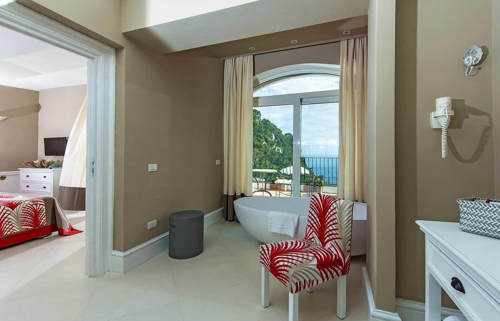 A hotel room with a bed, a chair, a small table, a trash bin, and a view of the outdoors.