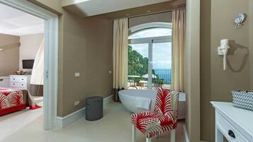 A hotel room with a bed, a chair, a small table, a trash bin, and a view of the outdoors.