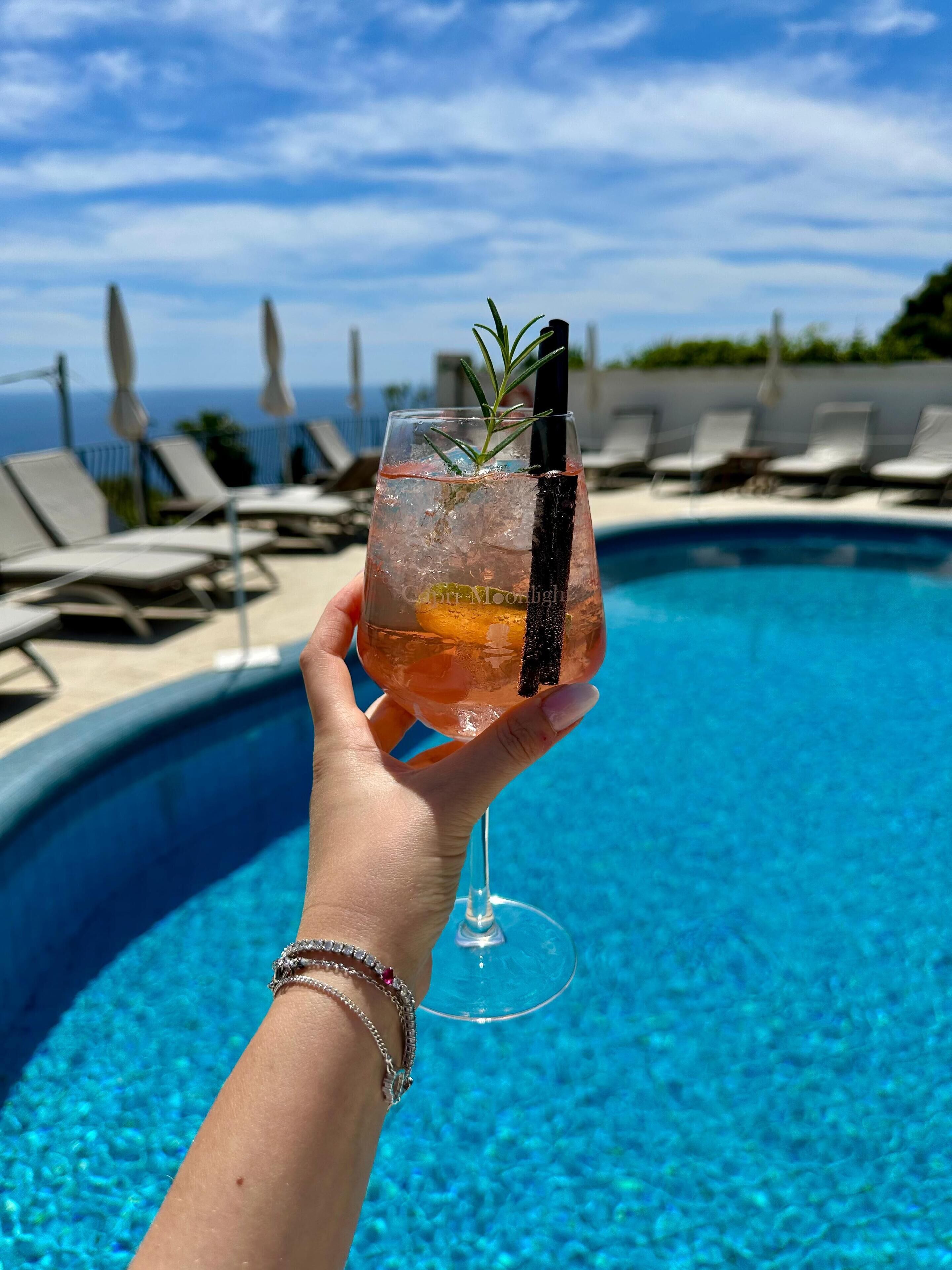 A hand holding a cocktail glass with a garnish, over a pool with lounge chairs and a view of the ocean.