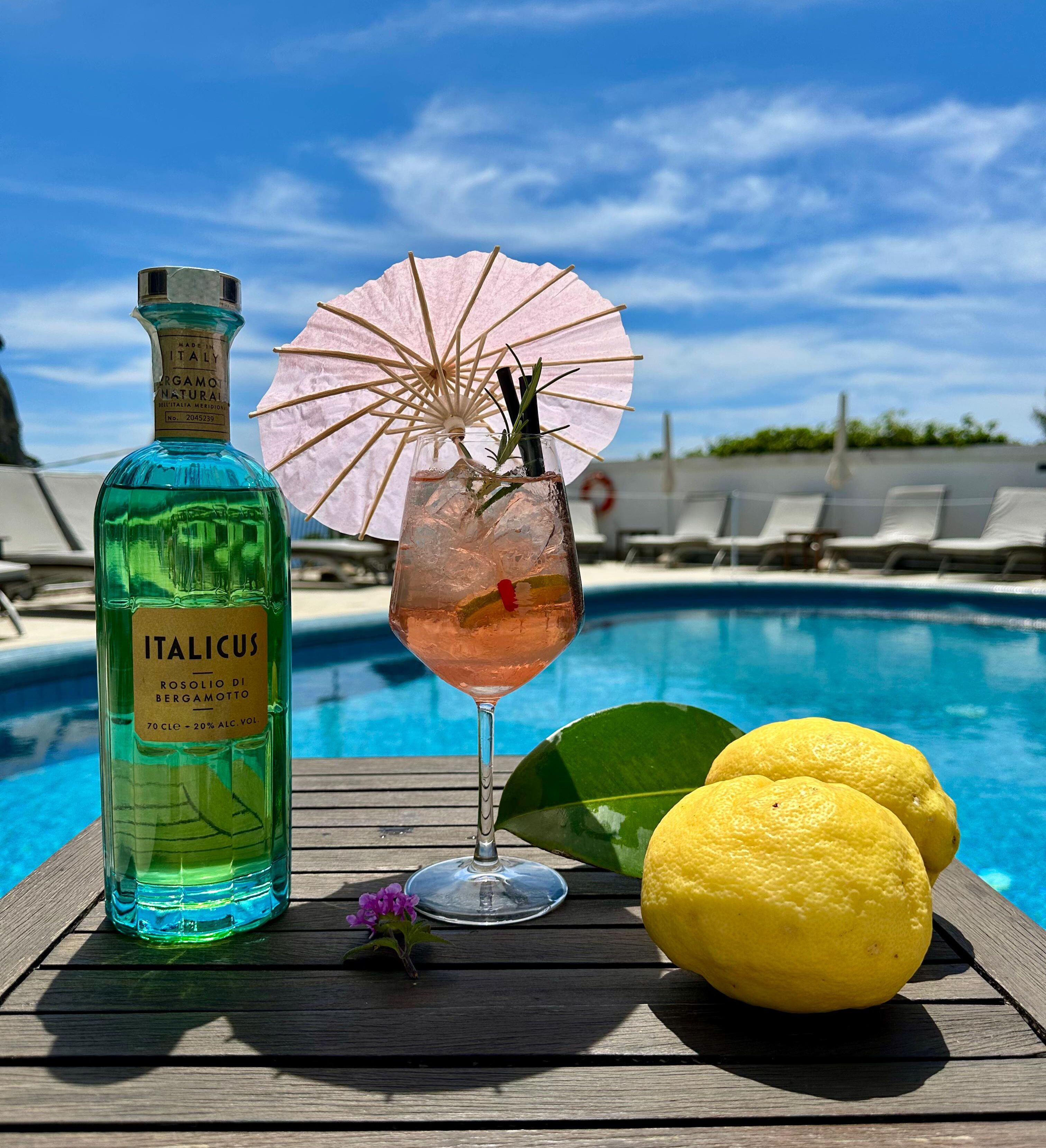 A bottle of ITALICUS rosolio di bergamotto, a glass of rosé cocktail with a pink umbrella, and lemons on a wooden table by a pool.