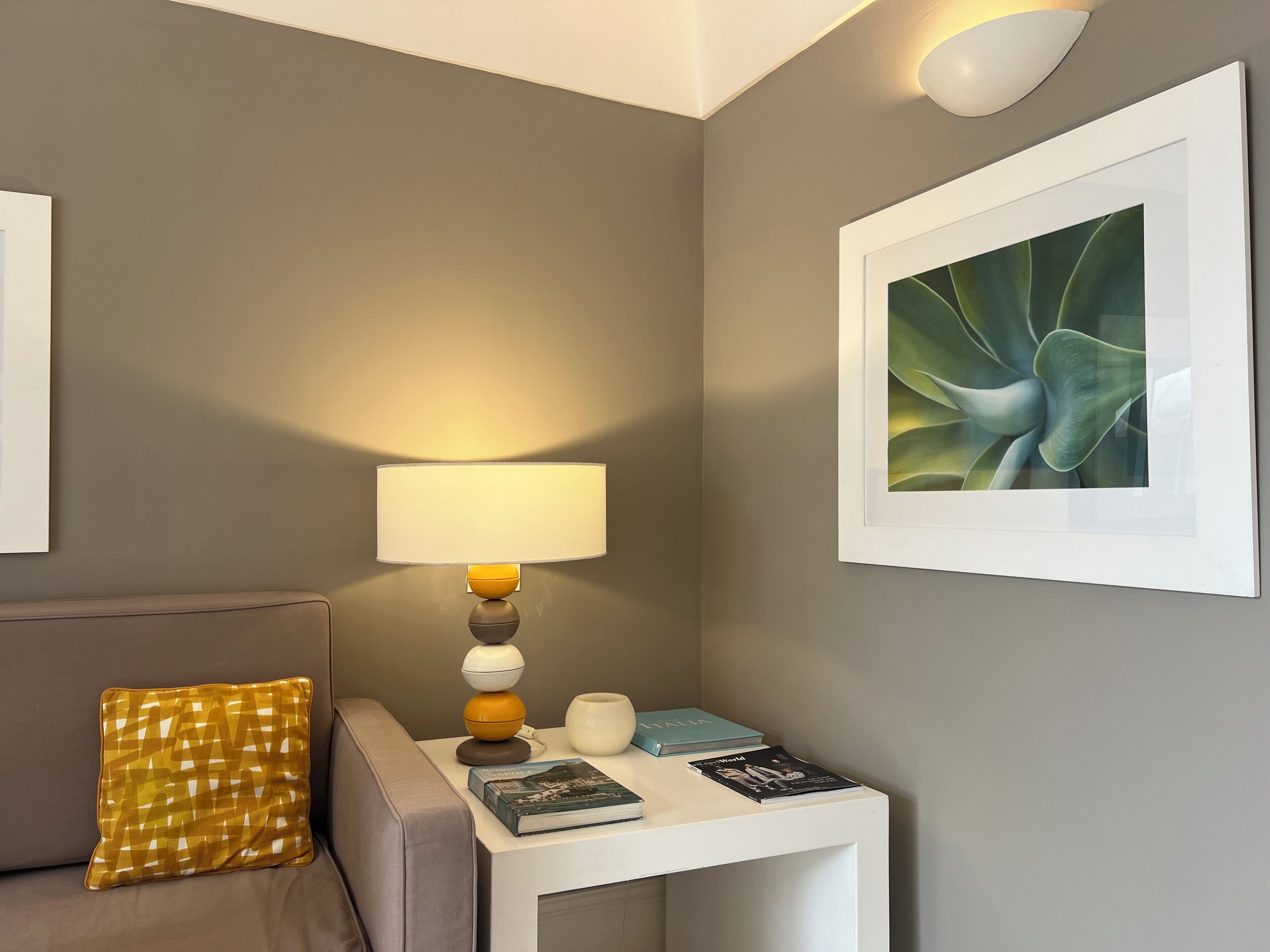 Una habitación moderna con pared gris, una mesita de noche blanca, una lámpara, un cojín y una obra de arte enmarcada.