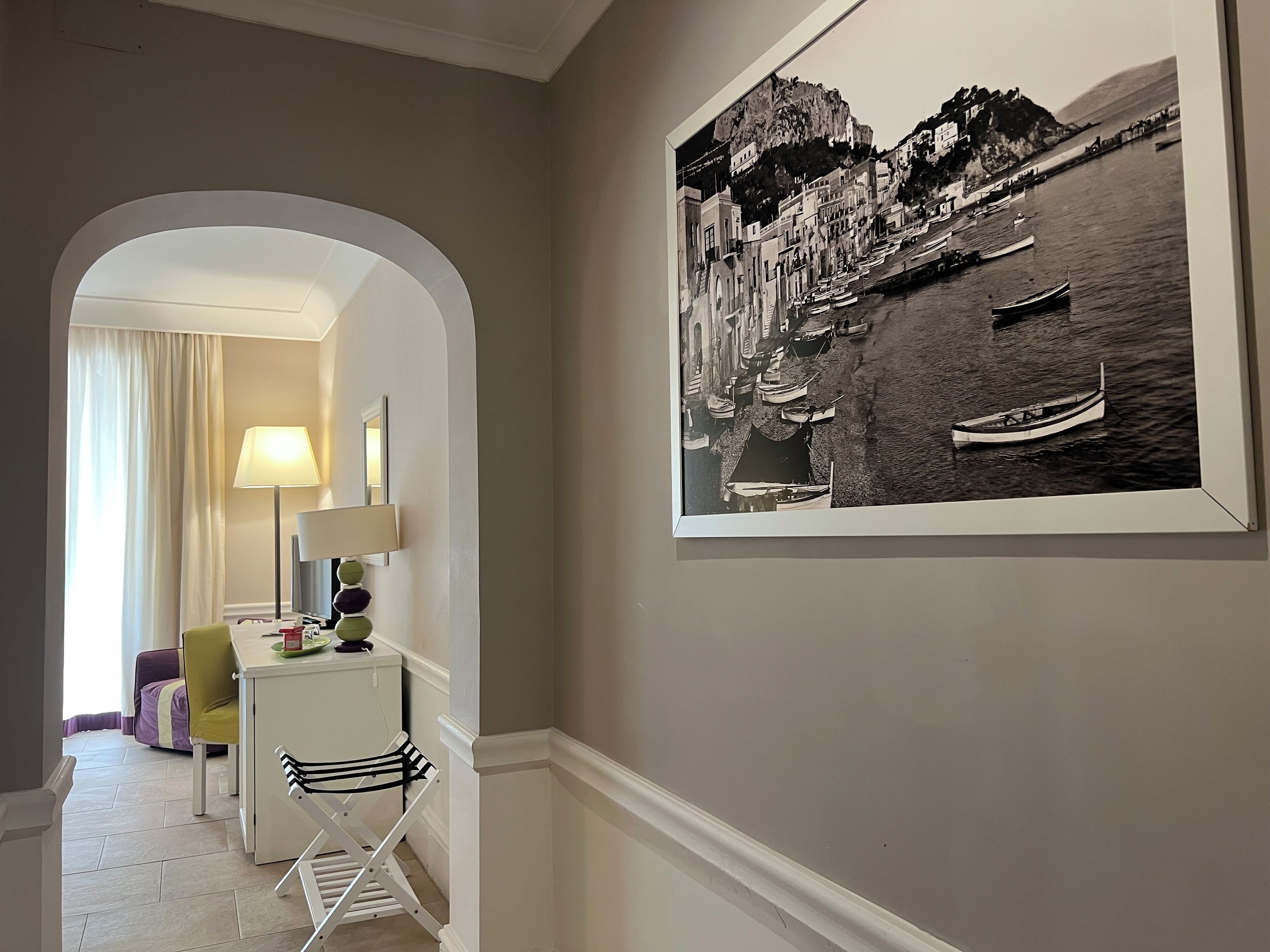 A hallway with a framed black and white photograph of a coastal town, a desk with a lamp, and a chair.