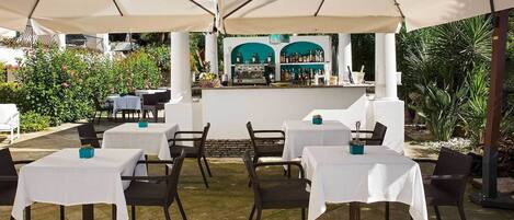 An outdoor patio with tables and chairs, a bar area, and lush greenery in the background.