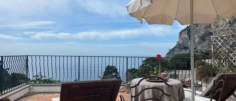 A terrace with outdoor seating, a table with a white umbrella, and a view of the sea and cliffs.