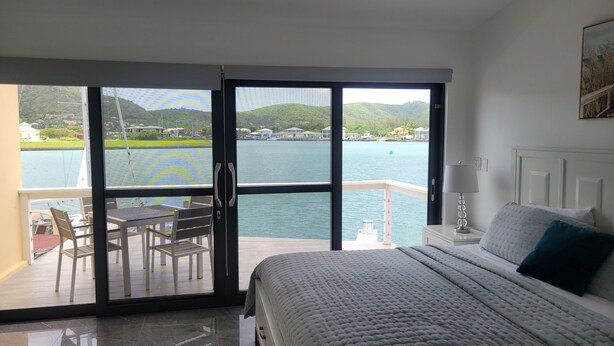 A bedroom with a bed, a nightstand, a lamp, and a view of a body of water through a sliding glass door. - HBK Villa Rentals at Jolly Harbour (Jolly Harbour)