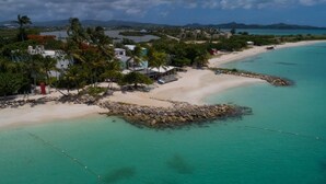An aerial view of a coastal area with a sandy beach, clear turquoise waters, and a rocky breakwater. - HBK Villa Rentals at Jolly Harbour (Jolly Harbour)