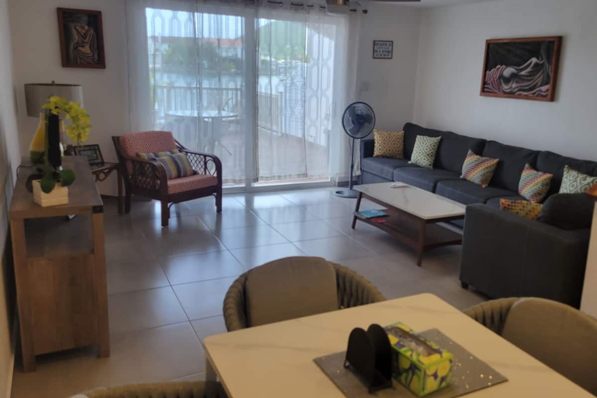 A living room with a sofa, armchairs, a coffee table, and a dining table. There is a fan, a plant, and artwork on the walls.