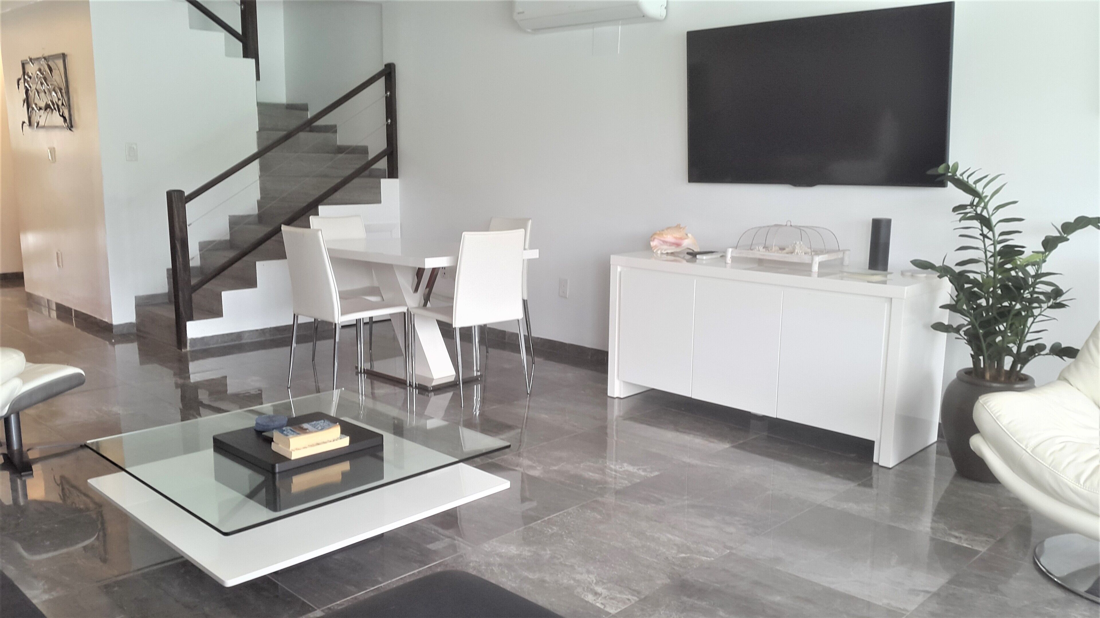 A modern living room with a glass coffee table, white furniture, a staircase, and a flat-screen TV mounted on the wall.