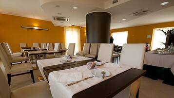 A hotel dining area with multiple tables and chairs, a central pillar, and a view of the room.