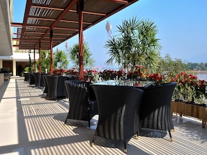 A covered outdoor seating area with wicker chairs and tables, surrounded by potted plants and flowers, overlooking a body of water.