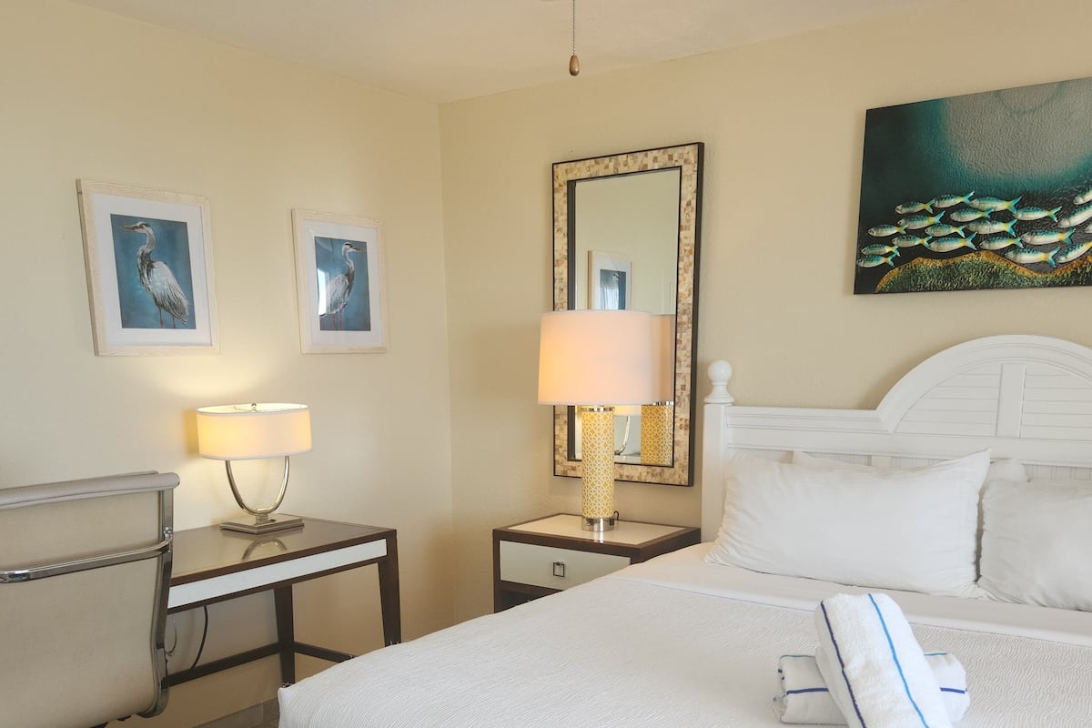 A hotel room with a bed, a desk, a chair, a lamp, a mirror, and artwork on the walls.