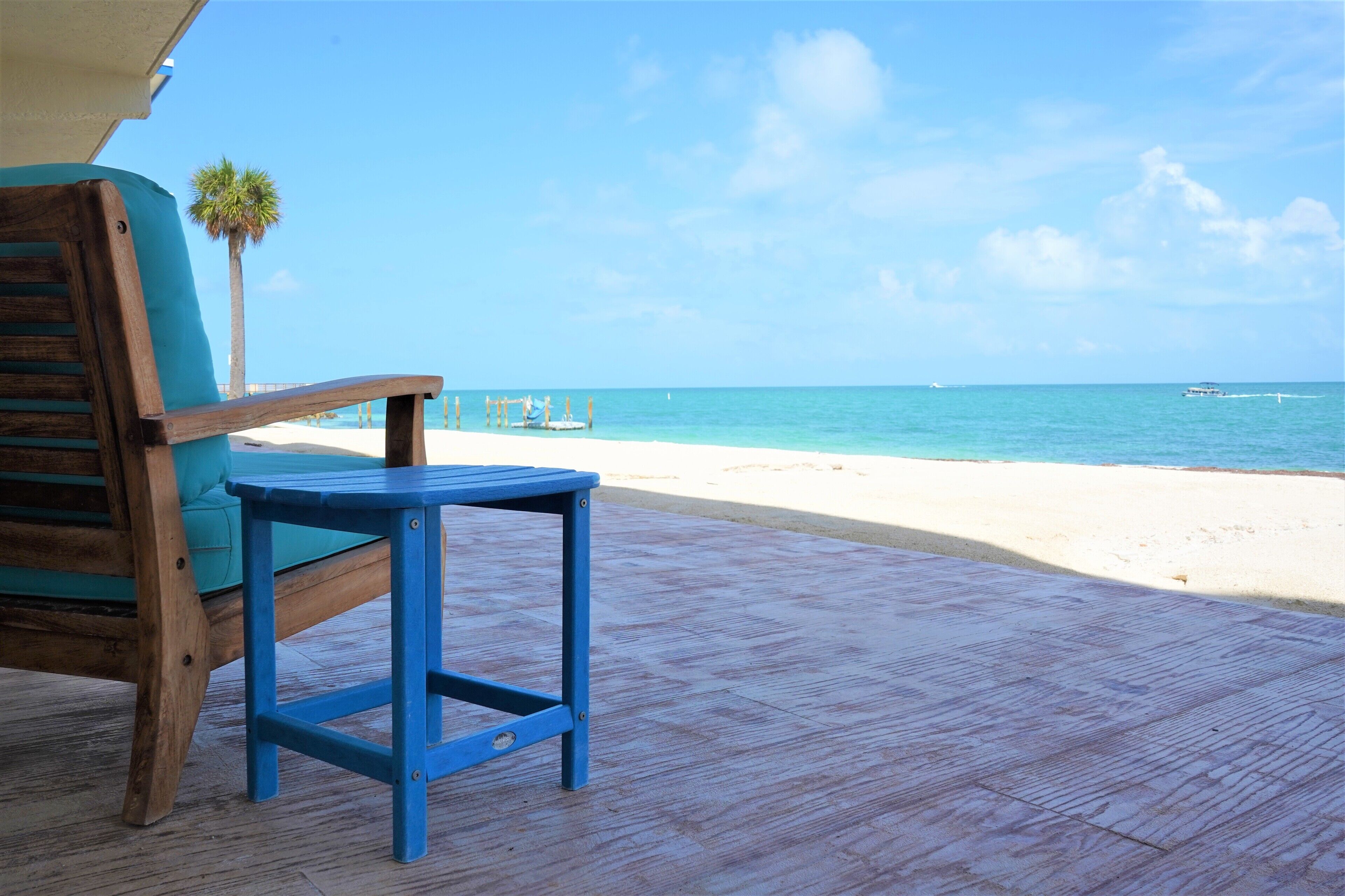 A wooden chair and table on a wooden deck overlooking a beach with clear blue water and a palm tree.