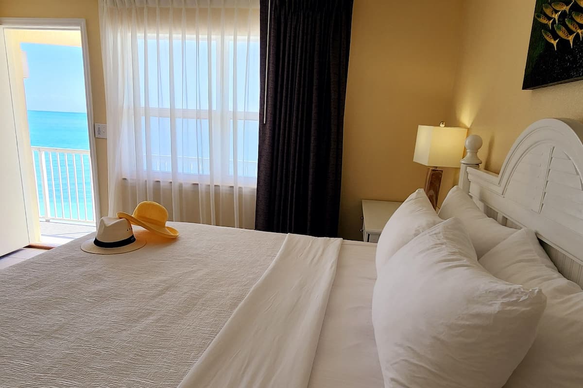 A hotel room with a bed, a nightstand with a lamp, a window with curtains, and a view of the ocean.