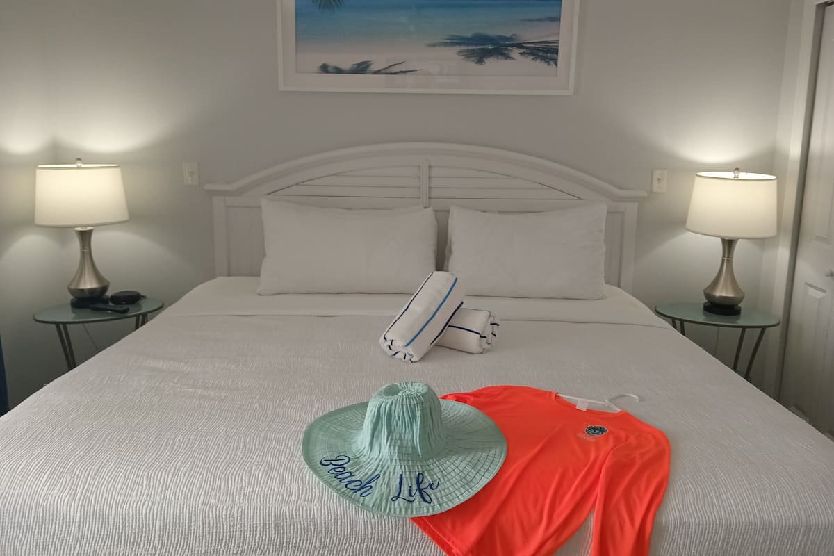 A neatly made bed with white linens, a framed picture of a beach, two bedside lamps, and a small table with a hat and a red garment on it.