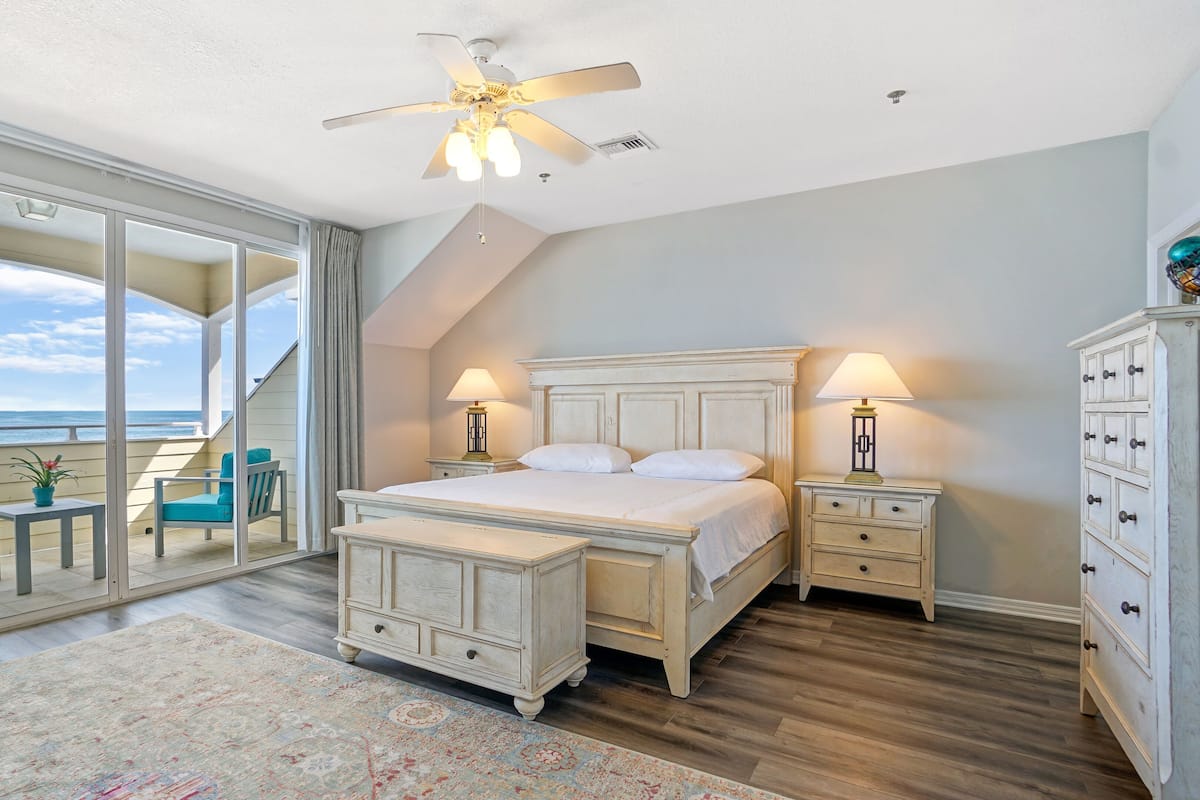 A bedroom with a large bed, two bedside tables with lamps, a dresser, and a view of the ocean through a sliding glass door.