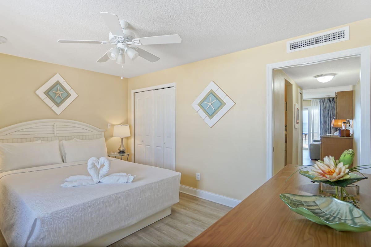 A hotel room with a bed, two bedside lamps, a ceiling fan, and a view of the ocean through a door.