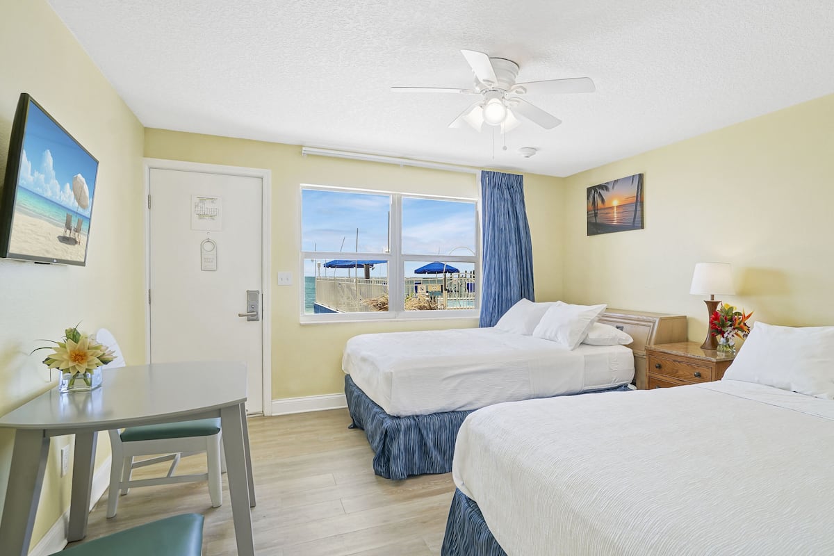 A hotel room with two beds, a desk, a window with a view of the beach, and a ceiling fan.