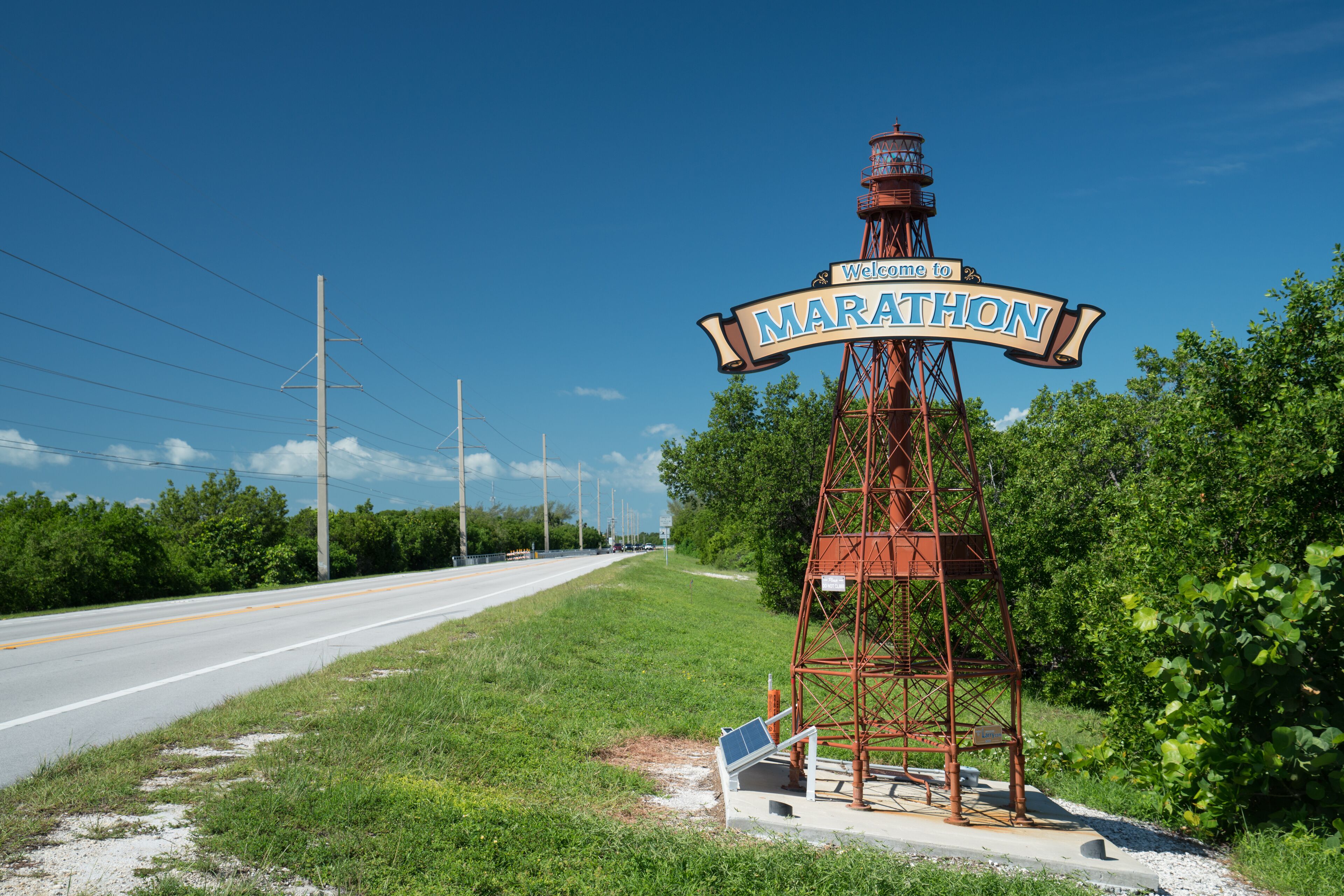 A signpost with "Welcome to Marathon" written on it, located beside a road with greenery on the sides.