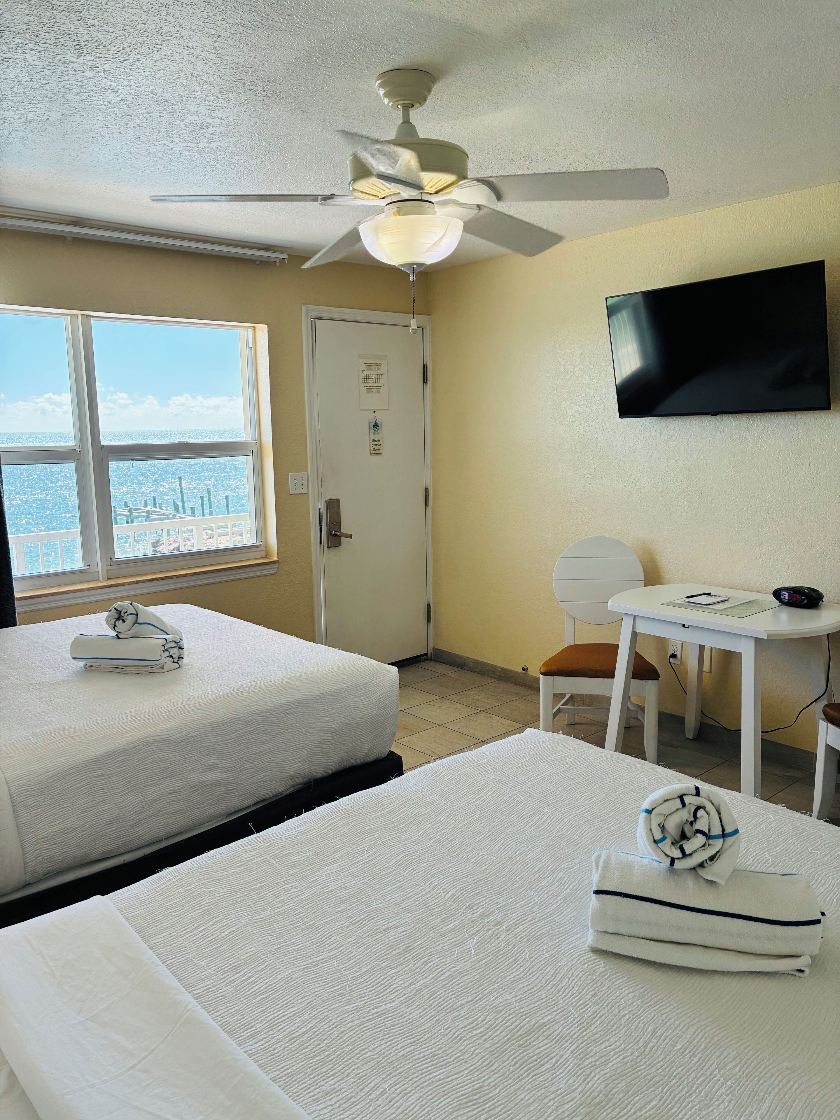Habitación de hotel con dos camas, ventilador de techo, televisor, escritorio con silla y ventana con vista a la ciudad.