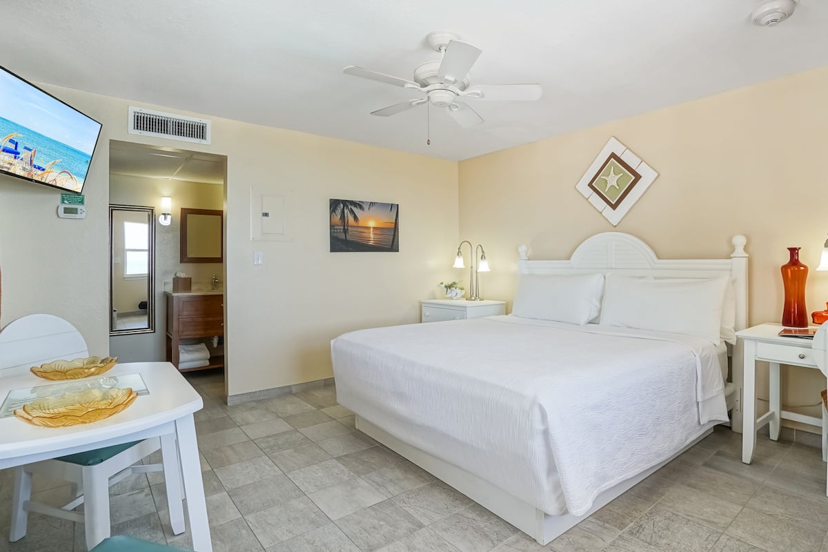 A hotel room with a bed, bedside tables, a desk, a chair, a ceiling fan, a TV, and a bathroom visible through an open door.
