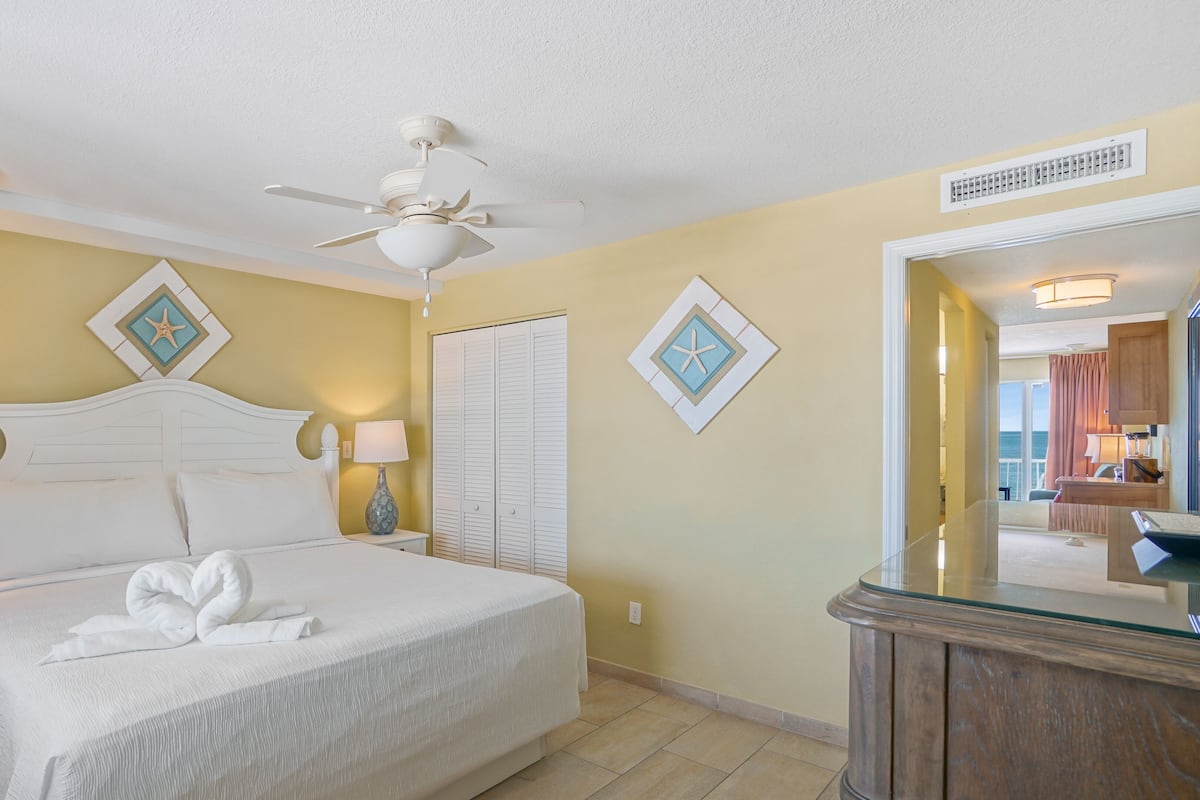 A hotel room with a bed, two bedside lamps, a ceiling fan, a wardrobe, and a bathroom mirror.