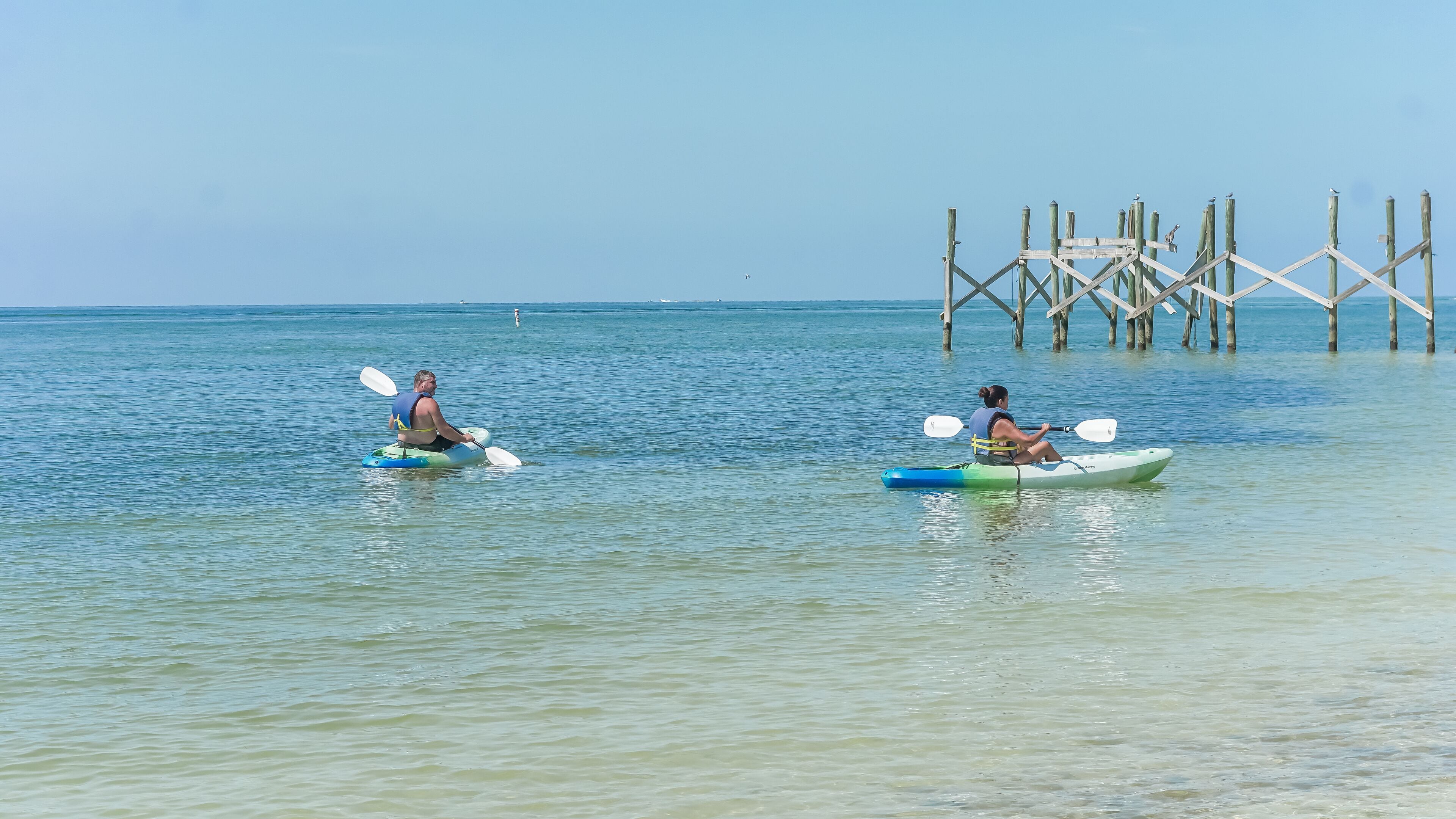 Deux personnes pratiquant le kayak sur une mer calme, avec une jetée en bois en arrière-plan.