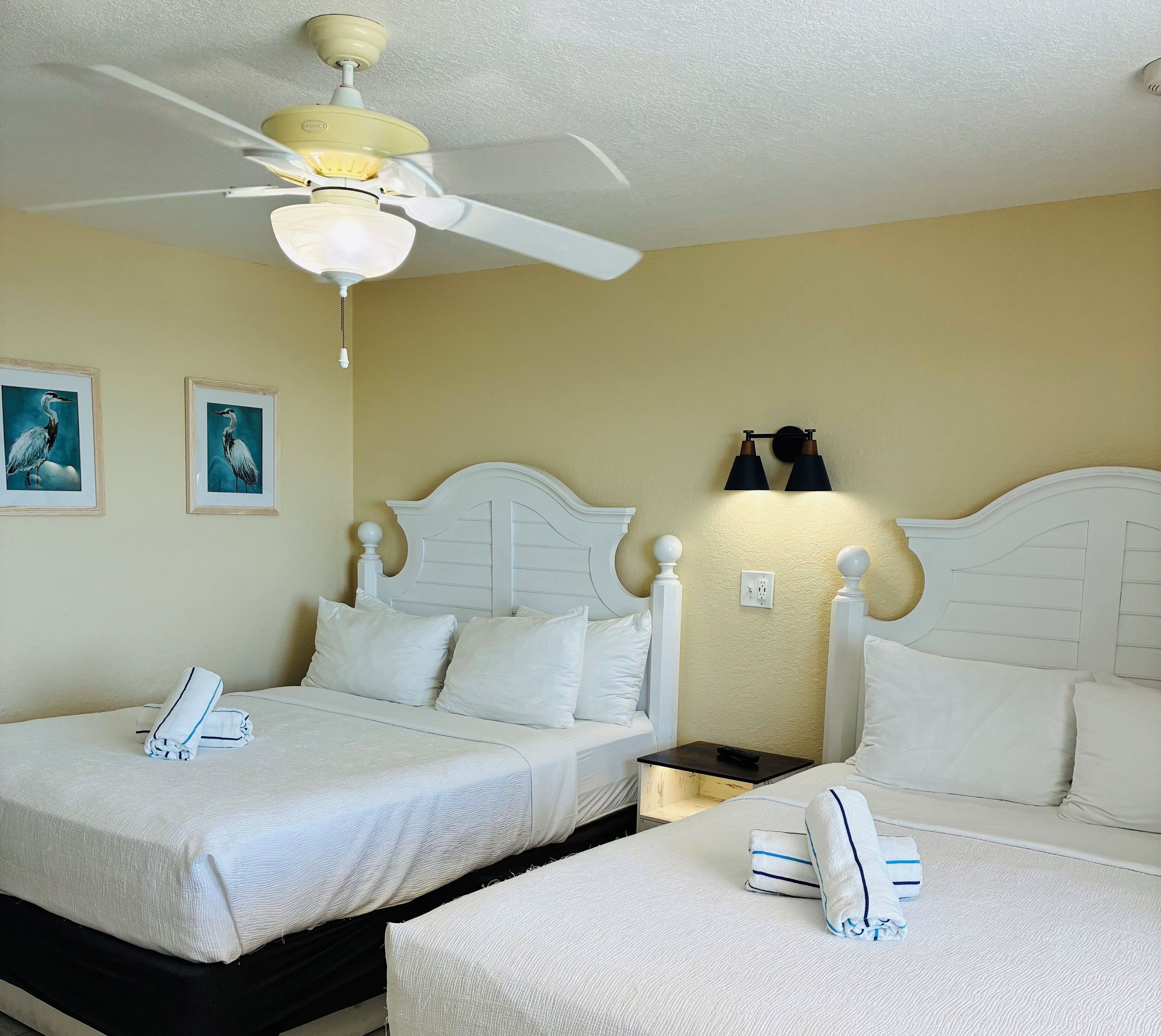 Une chambre d’hôtel avec deux lits, un ventilateur de plafond, des lampes fixées au mur et des œuvres d’art encadrées.