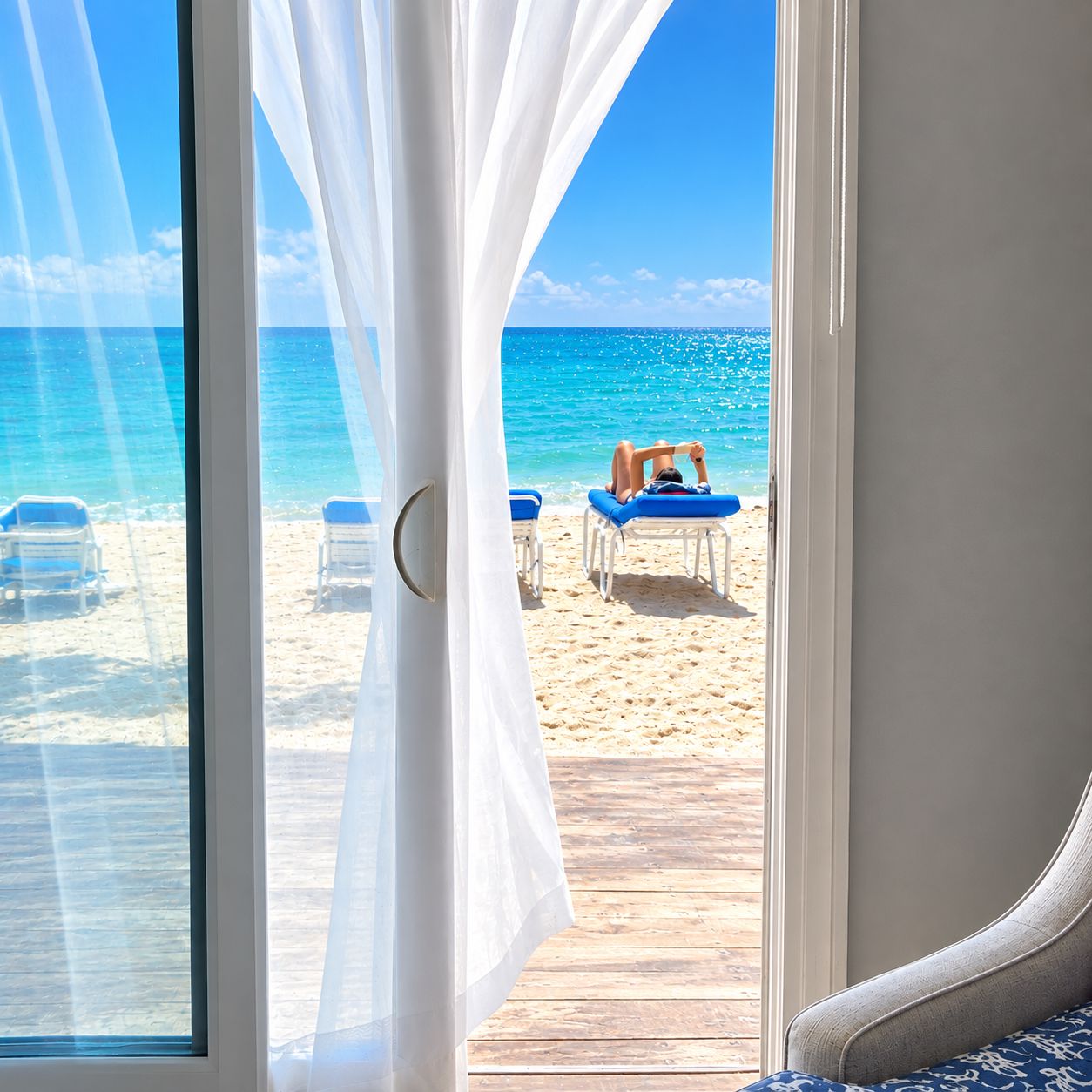 A beach view through a window with white curtains, a wooden deck, and beach chairs.