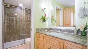 A bathroom with a glass-enclosed shower, a white toilet, a sink with a marble countertop, wooden cabinets, and a mirror with a towel hanging on the wall.