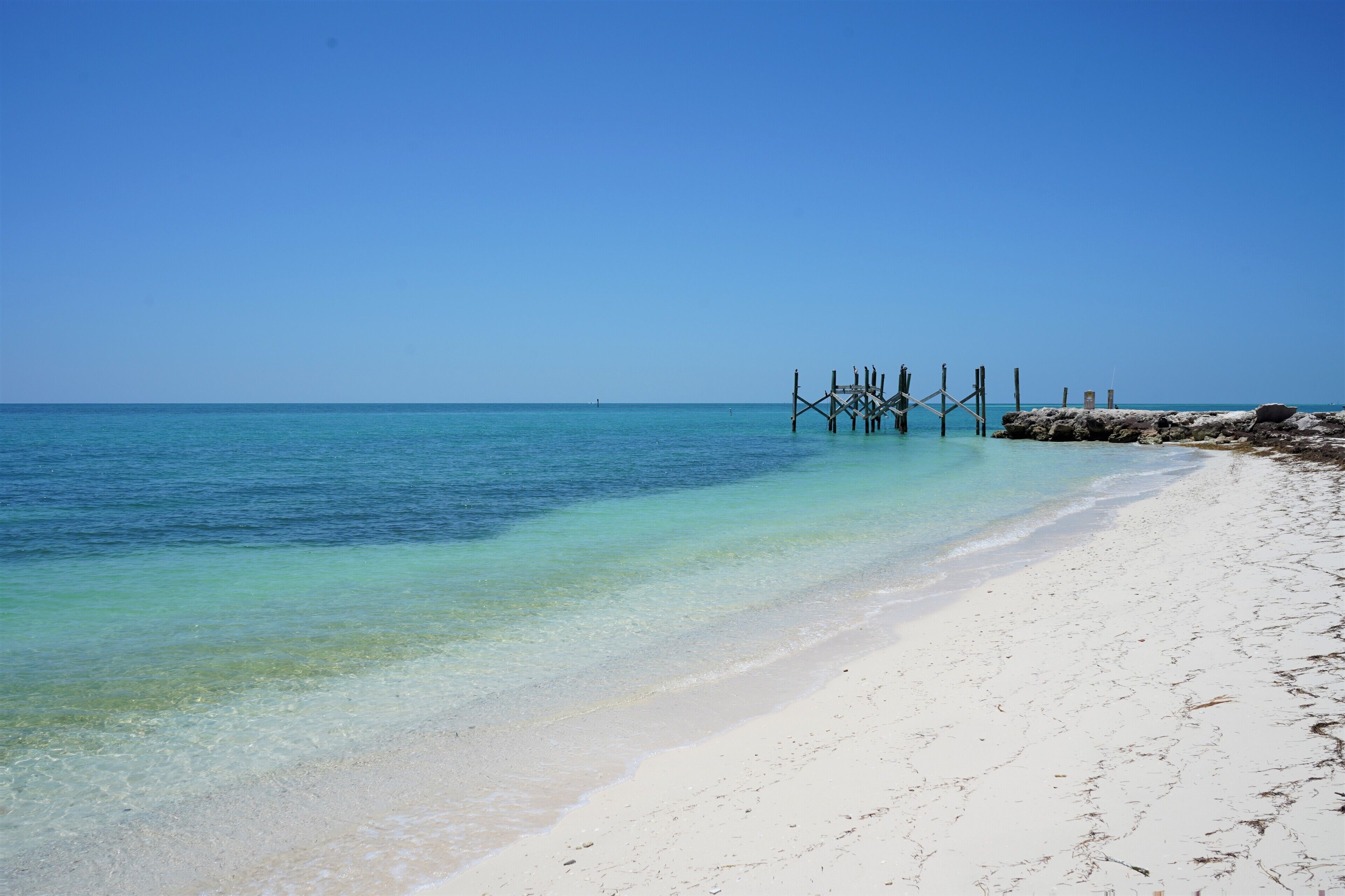 A beach with clear turquoise water, white sand, and a wooden pier structure.