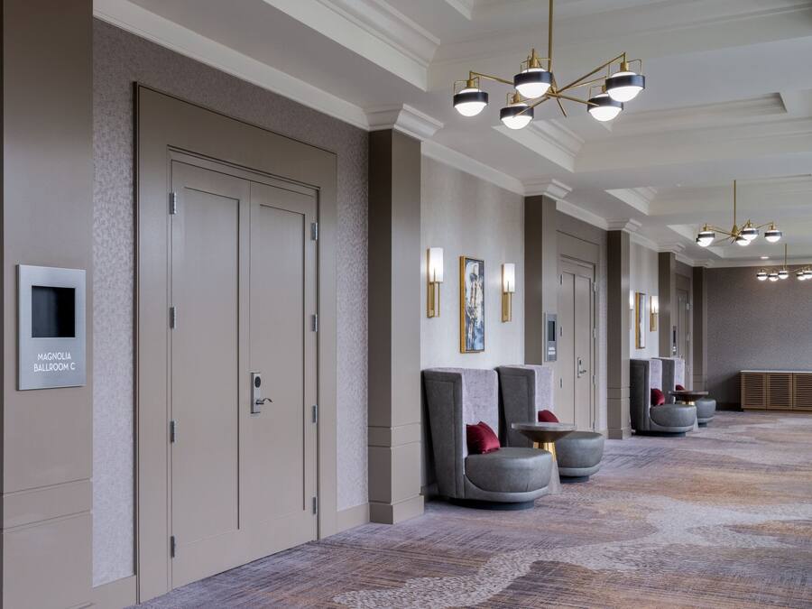 A hotel corridor with a carpeted floor, seating areas with cushions, and a ceiling with chandeliers.