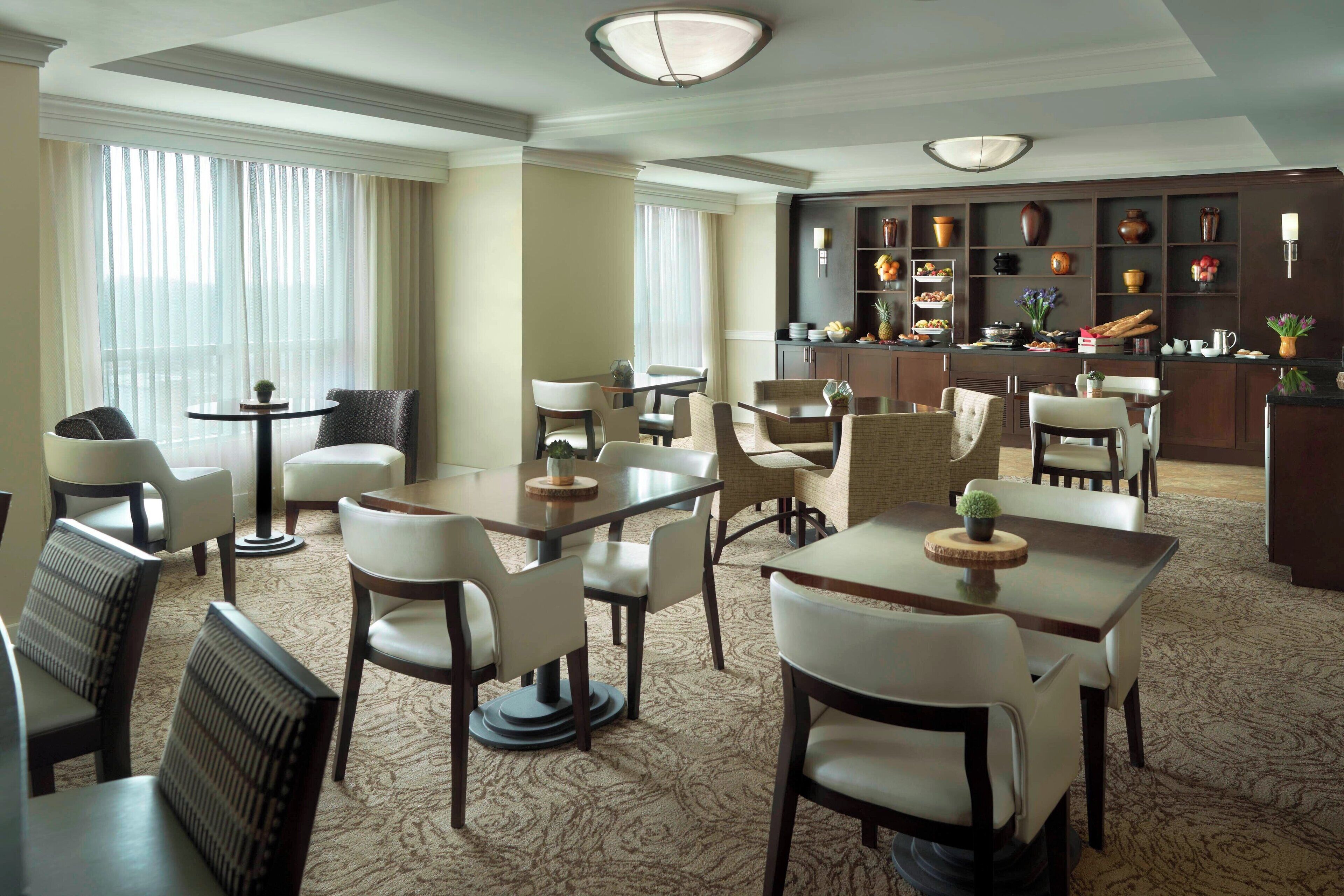 A hotel dining area with multiple tables and chairs, a bar area with shelves of decorative items, and a view of the city through large windows.