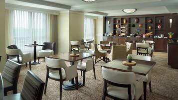 A hotel dining area with multiple tables and chairs, a bar area with shelves of decorative items, and a view of the city through large windows.