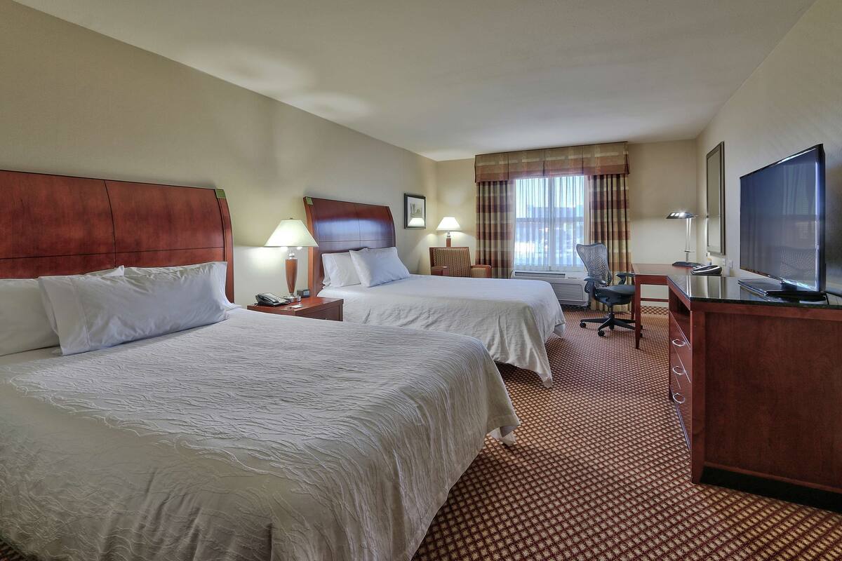 A hotel room with two beds, a desk, a chair, a TV, and a window with curtains.