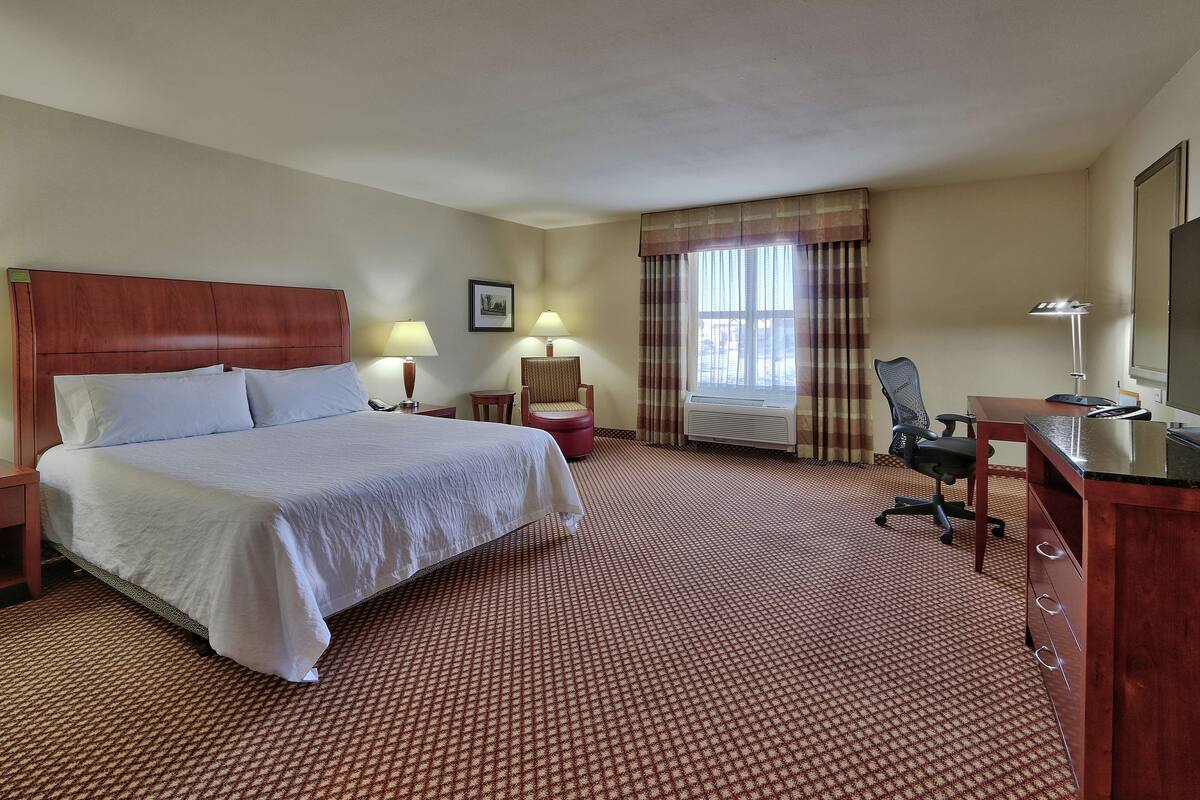 A hotel room with a large bed, two bedside tables with lamps, a desk with a chair, a television, and a window with curtains.