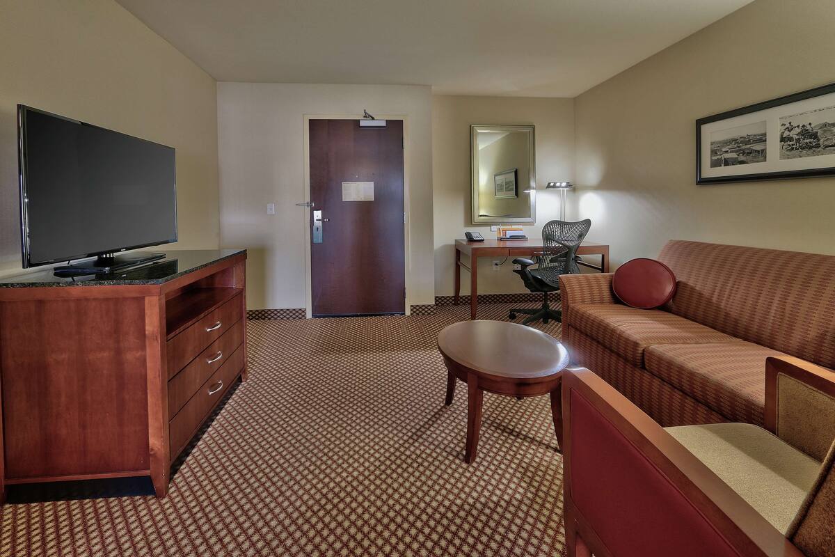 A hotel room with a flat-screen TV on a wooden stand, a wooden desk with a chair, a sofa, and a patterned carpet.
