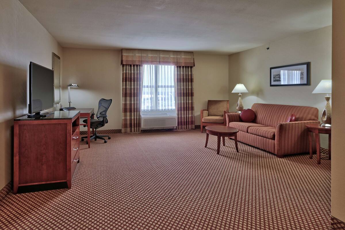A hotel room with a red carpet, a desk with a computer, a TV, a sofa, a chair, and a window with curtains.