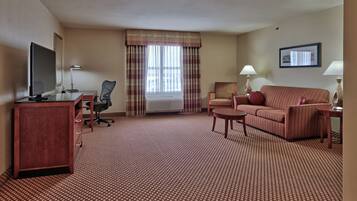 A hotel room with a red carpet, a desk with a computer, a TV, a sofa, a chair, and a window with curtains.