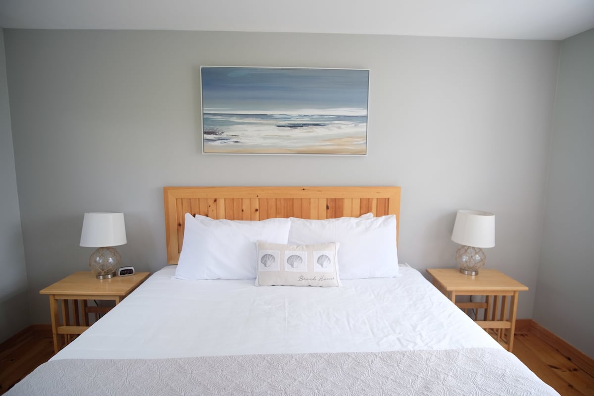 A neatly made bed with white linens, a wooden headboard, and a beach-themed pillow. There are two bedside tables with lamps and a painting of a beach scene above the bed.
