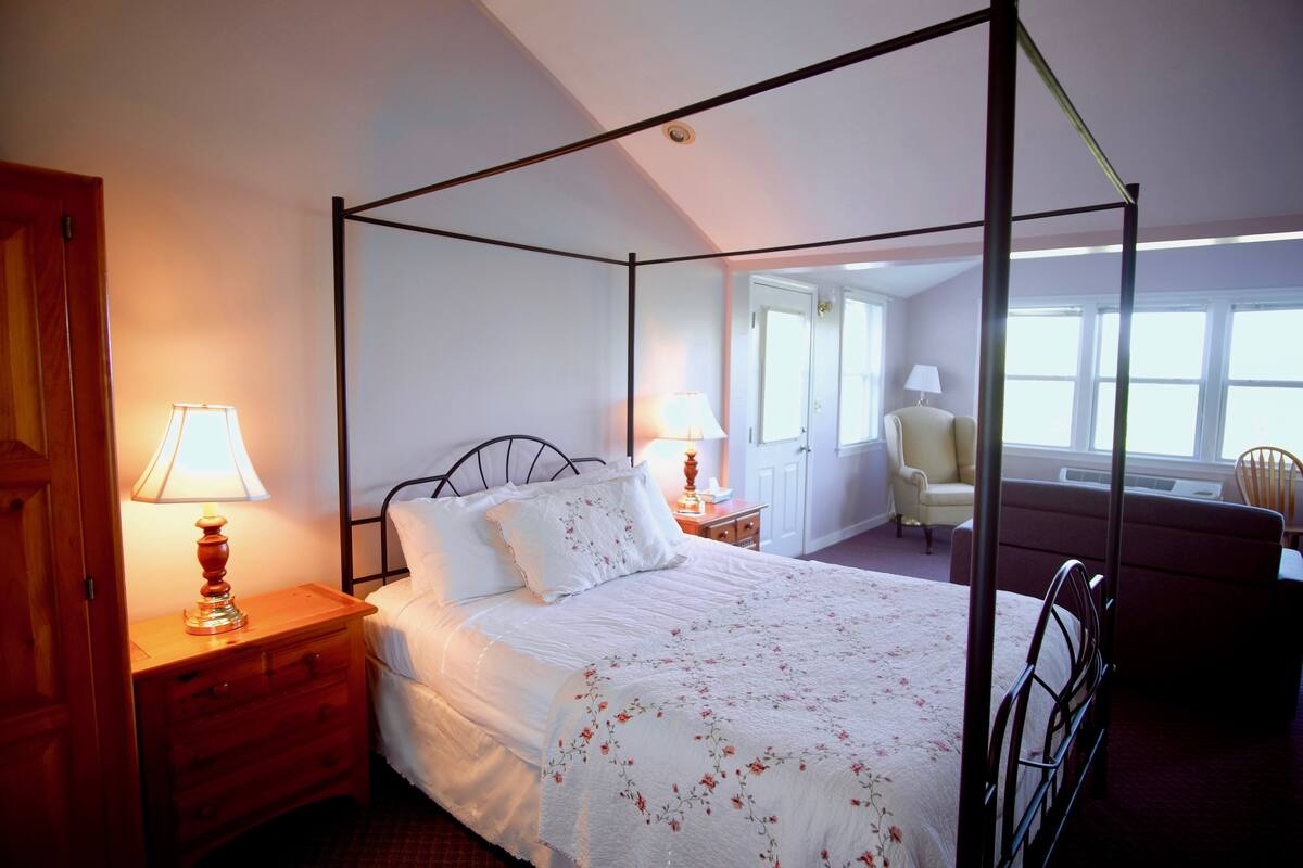 A four-poster bed with a white bedspread and floral pattern, flanked by wooden nightstands with lamps, in a room with a view of a chair and a window.