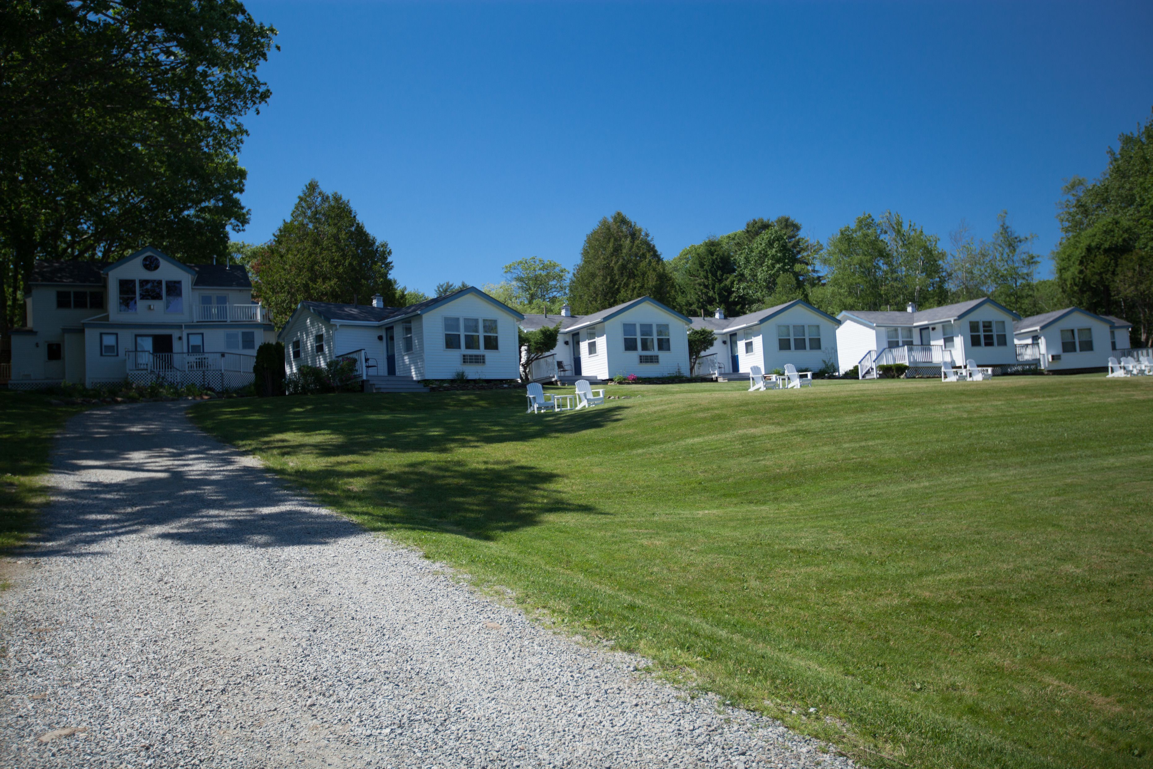 A row of white cabins with a gravel path leading to them, surrounded by a grassy area and trees.