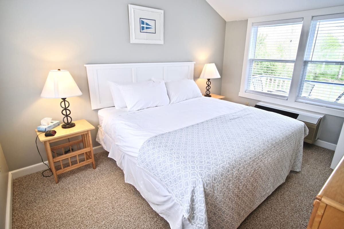 A neatly arranged bedroom with a large bed, two bedside lamps, a small wooden nightstand, and a window with blinds.