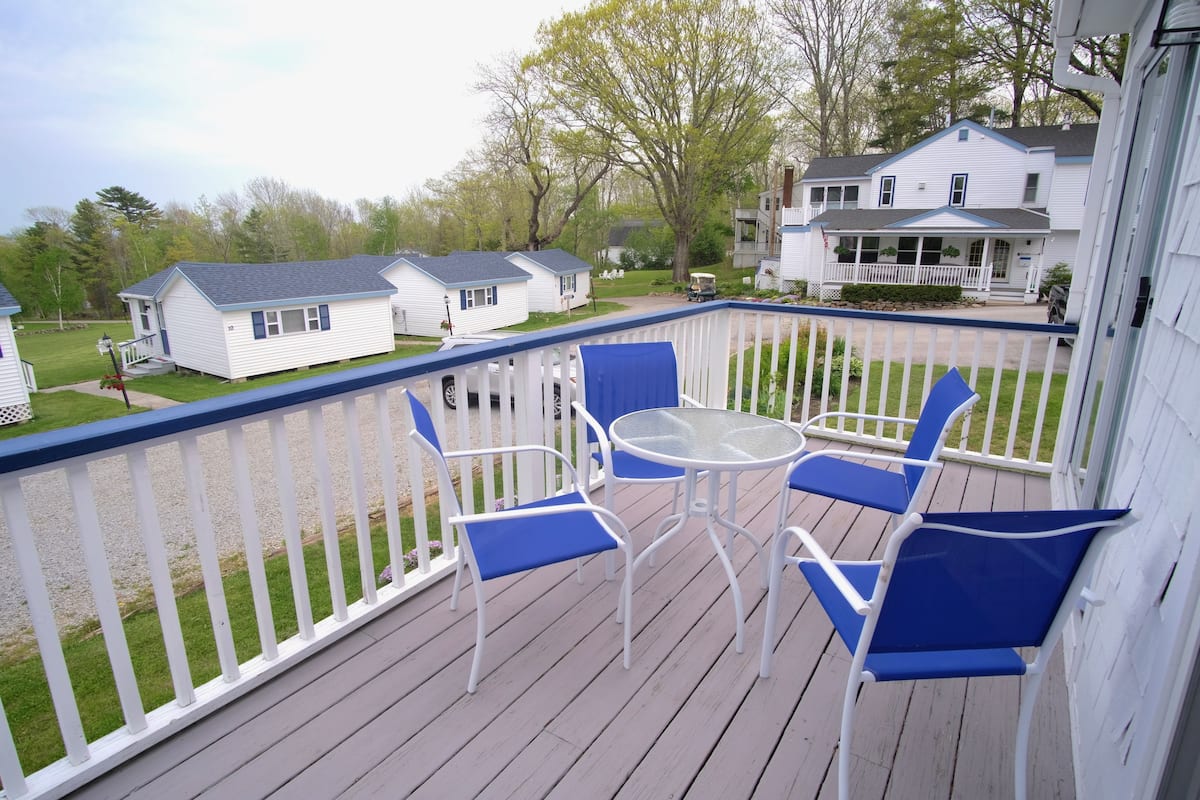 A covered outdoor deck with a table and four blue chairs, overlooking a residential area with houses and a golf cart.