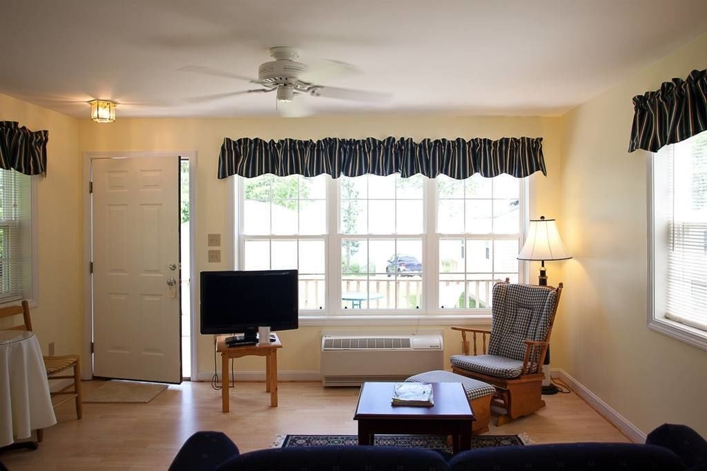 Ein Wohnzimmer mit Fernseher, Deckenventilator, einem Fenster mit Vorhängen, einem Sessel und einem Tisch.