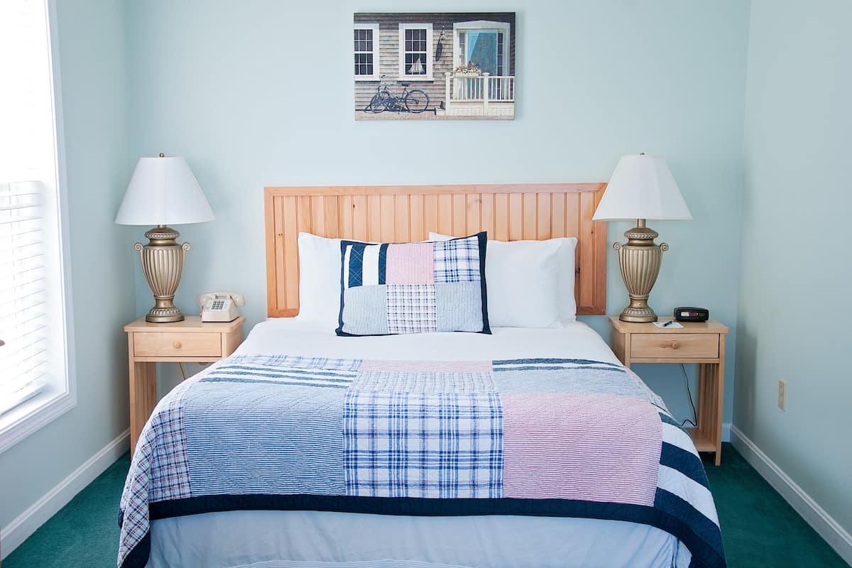 A neatly made bed with a blue and white checkered bedspread, flanked by two bedside tables with lamps and a small clock. A framed picture hangs above the bed.